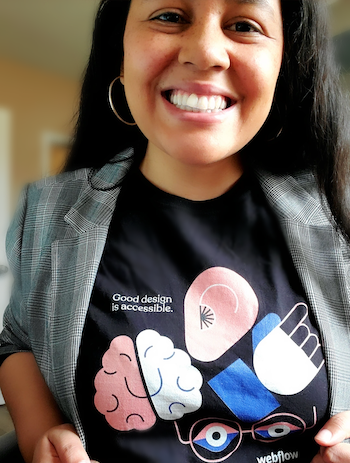 Deyra Jaye Fontaine smiles at the camera proudly while showing off her t-shirt that reads, "Good design is accessible". The shirt has symbols to represent neurodiverse, hearing impaired, vision impaired and mobility impaired peoples.
