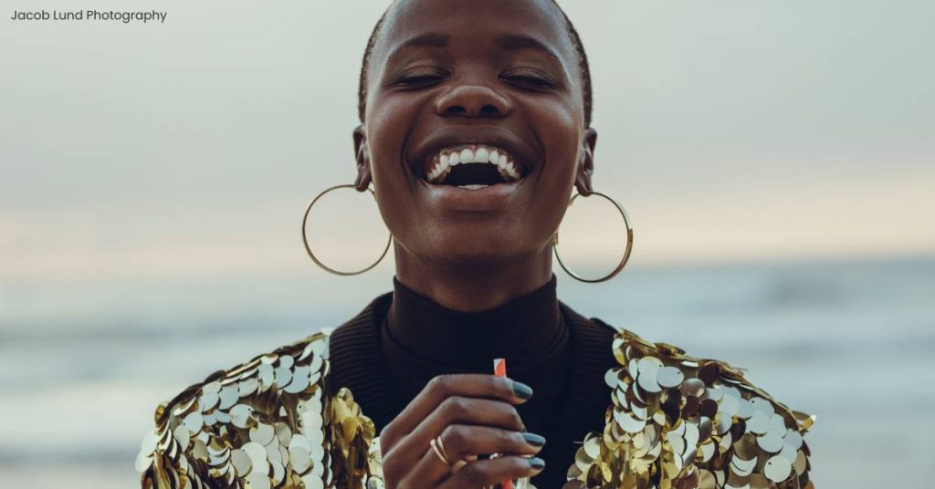 A dark-skinned woman is laughing as she stands in front of the ocean sipping on a soda. She is wearing gold hoop earrings and a gold sequin blouse. She is bald and her nails are painted navy blue.