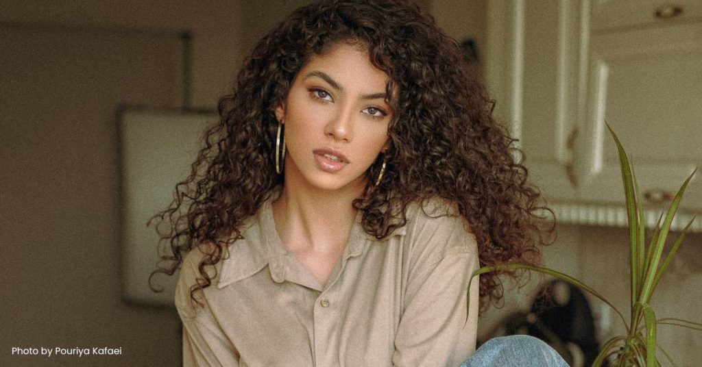 A racially ambiguous model sitting in a photography studio. She has light skin, brown curly hair, hazel eyes and full lips.
